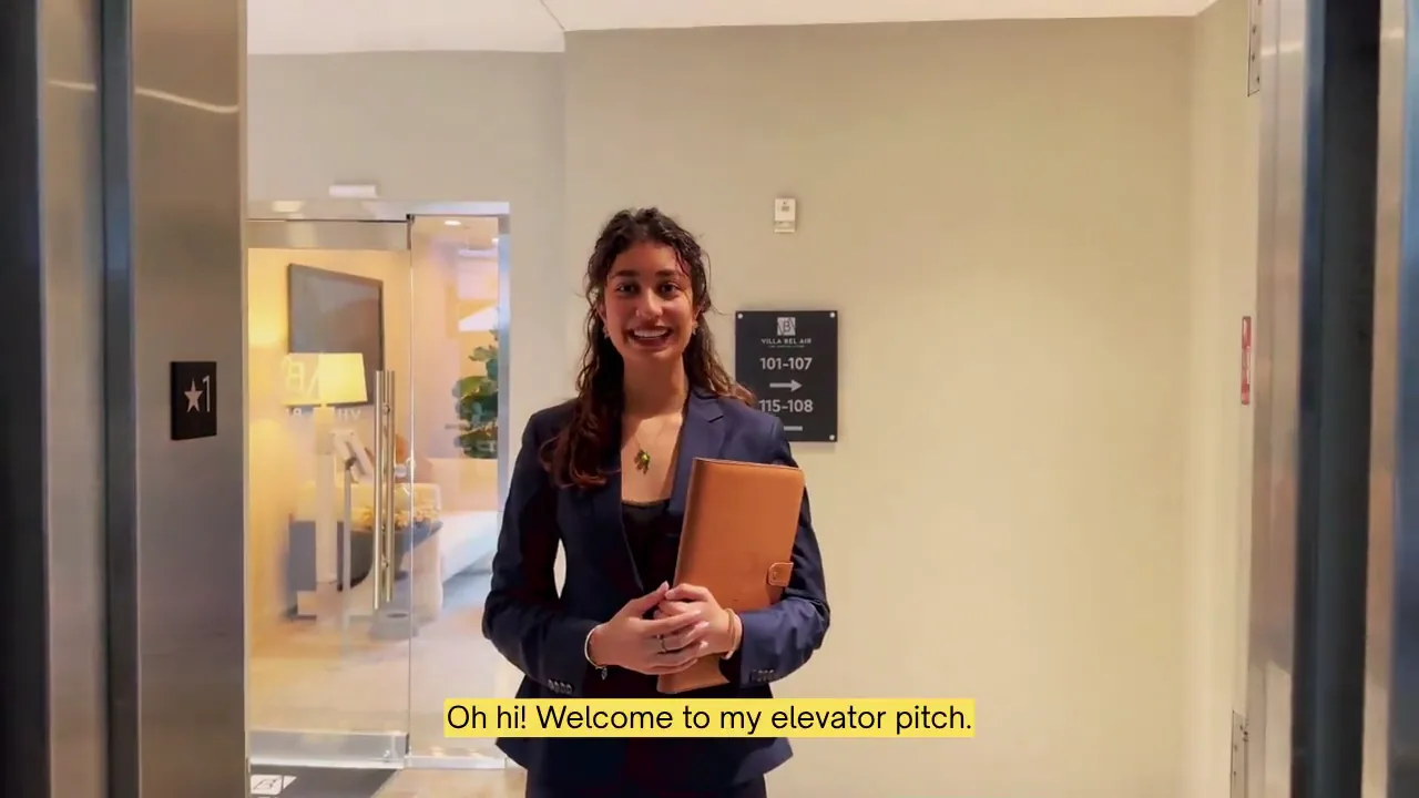 Aditi smiling by her building's elevator, holding a leather folio, as her elevator pitch begins.